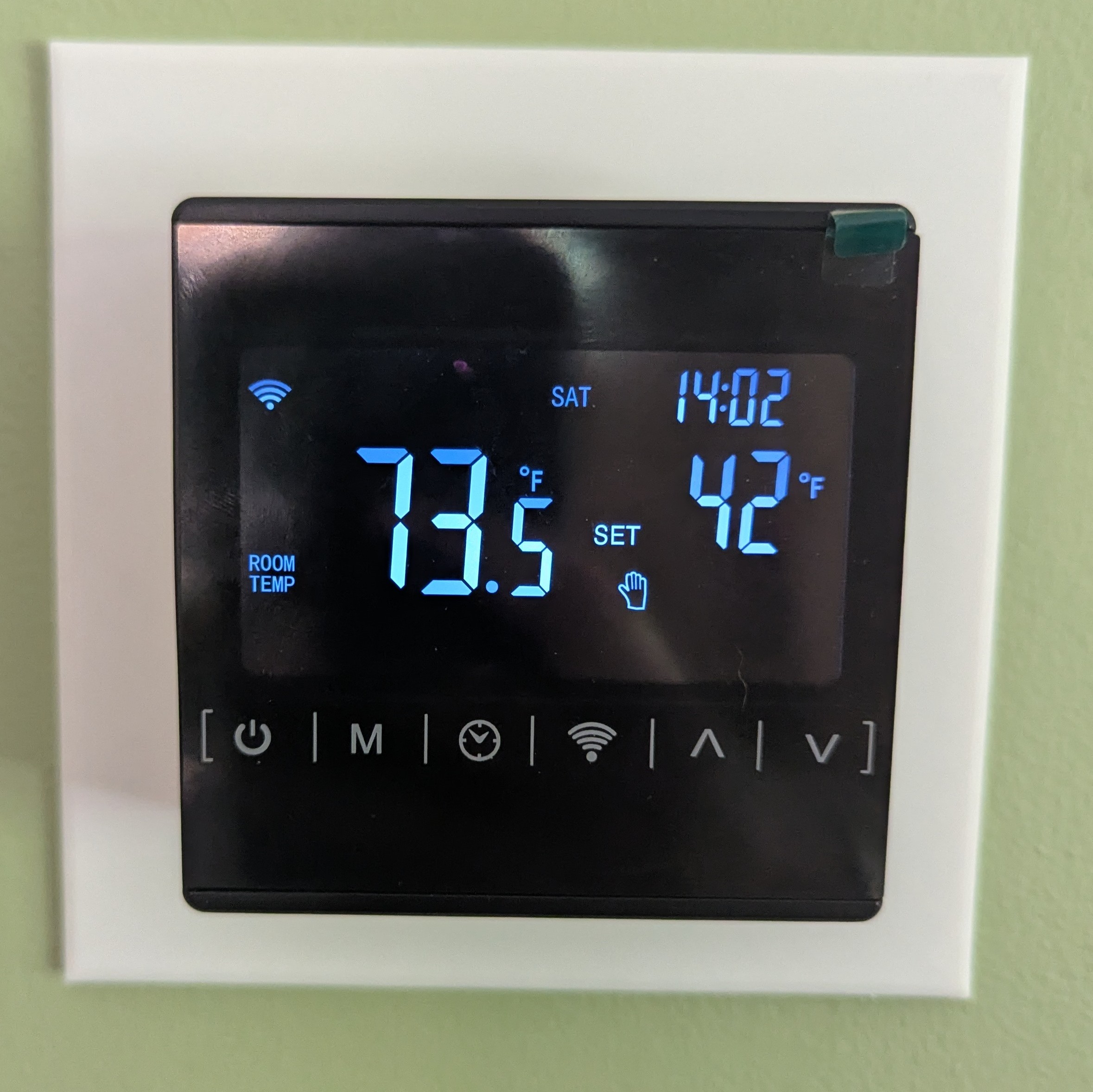 A photograph of a green wall with a black digital thermostat and a white square around the thermostat. The screen reads 73.5° Fahrenheit. with the set point of 42° Fahrenheit